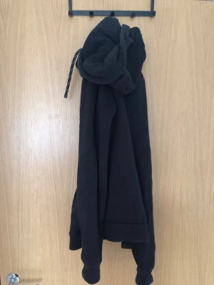 Svart hoodie med regnbågsmärke - Svart hoodie med huva och dragsnören, samt en liten broderad regnbågsfärgad detalj på bröstet. Klassisk känguruficka framtill och ribbade muddar vid ärmslut och nederkant. Perfekt för en chill och avslappnad stil.