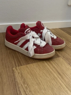 Röda Adidas Campus sneakers - Säljer ett par röda Adidas Campus sneakers med klassiska vita ränder och breda vita skosnören. Skorna har låg profil och är tillverkade i mocka med gummisula. Adidas-logga på plösen och sidan. Perfekta för dig som gillar retrostil och streetwear.