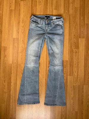 Ljusblå bootcut jeans från True Religion - Snygga ljusblå bootcut jeans från True Religion med klassiska fem fickor och dekorativa sömmar. Jeansen har låg midja och utsvängda ben, samt detaljerade bakfickor med knappar. Perfekta för en avslappnad och trendig look.