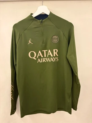 Grön PSG träningströja Jordan - Snygg grön långärmad träningströja från Jordan x PSG med halv dragkedja, tryckt Qatar Airways-logga och Paris Saint-Germain-emblem på bröstet. Tillverkad i funktionsmaterial som andas, perfekt för fotbollsträning eller gym. 