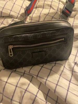 Svart axelväska från Gucci - Snygg svart axelväska från Gucci med klassiskt GG-mönster, silverfärgad dragkedja och justerbar axelrem i rött och blått. Väskan är rektangulär och har både läder- och textildetaljer. Perfekt för att bära det viktigaste med stil.