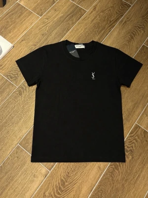Svart t-shirt från Saint Laurent - Svart t-shirt från Saint Laurent med klassisk passform och korta ärmar. Liten vit YSL-logga broderad på bröstet. Tillverkad i mjuk bomull som känns skön mot huden. Perfekt för en stilren och enkel look.