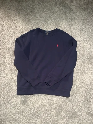 Mörkblå sweatshirt från Polo Ralph Lauren - Mörkblå sweatshirt från Polo Ralph Lauren med klassisk rund halsringning och lång ärm. Tröjan har en liten röd broderad logga på bröstet och är tillverkad i mjukt bomullsmaterial. Perfekt för en avslappnad och stilren look. Storlek XL 18-20 på barn/ungdom sortimentet vilket ungefär motsvarar en lite bredare S