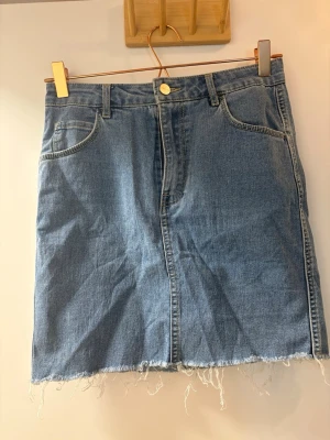 Blå jeanskjol med fransig kant - Snygg blå jeanskjol i klassisk modell med fem fickor och rå, fransig nederkant. Kjolen har normal passform och stängs med knapp och dragkedja framtill. Shell 79%, cotton 20% elastan 1%