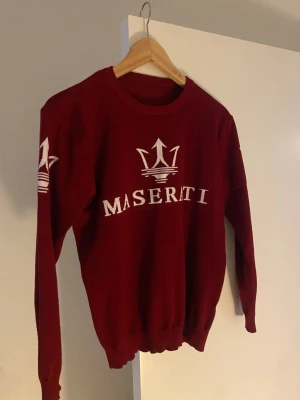 Vinröd Maserati stickad tröja - Snygg vinröd stickad tröja från Maserati med vit logga och text framtill samt på ärmarna. Tröjan har rund halsringning, långa ärmar och ribbade muddar. Perfekt för dig som gillar sportiga och exklusiva detaljer.