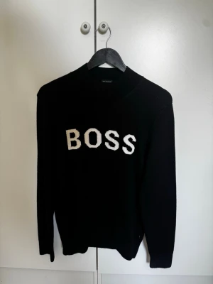 Svart stickad tröja från Hugo Boss - Snygg svart stickad tröja från Hugo Boss med vit BOSS-text framtill. Tillverkad av exklusivt italienskt garn. Tröjan har rund halsringning och långa ärmar, perfekt för dig som vill ha en stilren och trendig look.