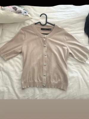 Beige kofta med kort ärm från Vero Moda - Söt beige kofta från Vero Moda med korta ärmar och dekorativa silvriga knappar formade som små rosetter. Koftan har rund halsringning och är stickad i ett mjukt material, perfekt för lager-på-lager och en clean look. Den är i bra skick och har använts max 3 gånger. Pris kan diskuteras.