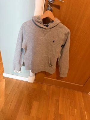 Grå hoodie från Polo Ralph Lauren - En stilren grå hoodie från Polo Ralph Lauren med klassisk huva och känguruficka framtill. 