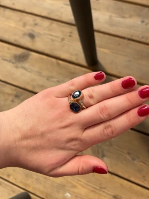 Ringar med blå sten och kristaller - Två eleganta guldfärgade ringar med stora blå stenar och små gnistrande kristaller runt den ena stenen. Ringarna har en klassisk och lyxig design som verkligen sticker ut. Perfekt för dig som gillar att bära unika accessoarer.