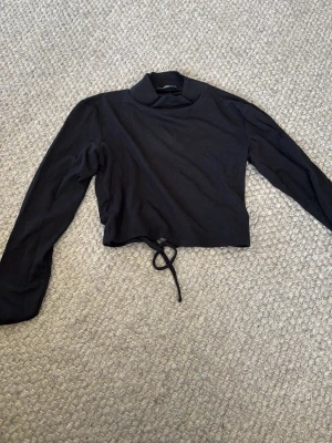 Svart croppad polotopp med snörning - Svart croppad polotopp med lång ärm och snörning framtill. Toppens material känns mjukt och stretchigt, perfekt för att skapa en cool och avslappnad look. Polokragen ger en trendig touch och snörningen gör att du kan justera passformen.