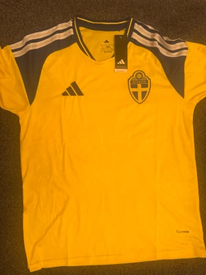 Sverige fotbollströja Adidas gul - Gul fotbollströja från Adidas med Sveriges landslagsemblem på bröstet och mörkblå detaljer på axlarna. Tröjan har subtila mönster i tyget och är tillverkad i ett lätt och ventilerande material, perfekt för träning eller match.