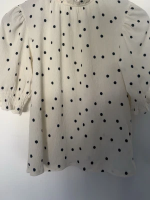 Prickig blus från Holly & Whyte - Vit blus med svarta prickar från Holly & Whyte by Lindex. Blusen har trekvartsärm med lätt puff och resår vid ärmslut. Tillverkad i ett krinklat tyg som ger en avslappnad känsla. Perfekt för dig som gillar klassiska mönster med en twist.