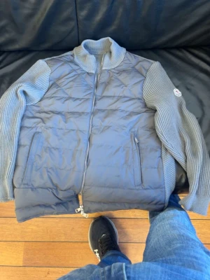 Grå pufferjacka från Moncler - Snygg grå pufferjacka från Moncler med stickade ärmar och krage. Jackan har quiltad front och rygg i glansigt material, två dragkedjefickor och Moncler-logga på ärmen. Perfekt för kalla dagar och riktigt trendig look.