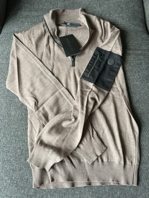 Beige tröja med dragkedja från Snoot - Stilren beige tröja från Snoot med halv dragkedja och svart ficka med dragkedja på ärmen. Tröjan har ribbade muddar och hög krage. Perfekt för en trendig och avslappnad look. Kan sänka priset vid en snabb affär!!