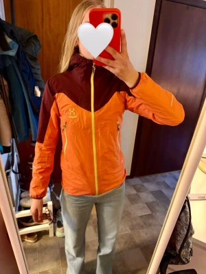 Orange och vinröd Haglöfs skaljacka - Snygg skaljacka från Haglöfs i orange och vinrött med gula dragkedjor. Jackan har hög krage, två sidofickor med dragkedja och är tillverkad i lätt, vattentätt material. Perfekt för utomhusaktiviteter och streetstyle. 