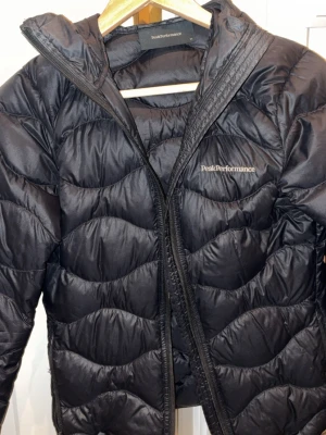 Svart dunjacka Peak Performance XS - Snygg svart dunjacka från Peak Performance i storlek XS. Jackan har vågformat quiltat mönster, dragkedja framtill och diskret logga på bröstet. Perfekt för kalla vinterdagar och riktigt lätt att bära. Klassisk modell som funkar till det mesta.