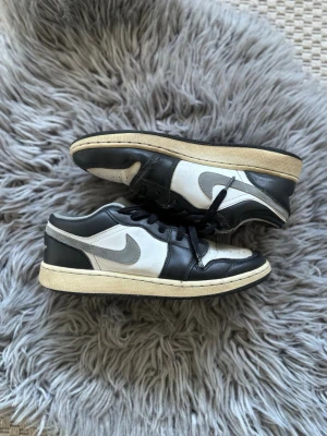 Nike Air Jordan 1 Low svart/vit - Säljer ett par Nike Air Jordan 1 Low sneakers i svart och vitt med grå swoosh och klassisk Air Jordan-logga på hälen. Skorna har lågt skaft, svarta skosnören och en beige sula. Tillverkade i läder för en clean och tidlös look.
