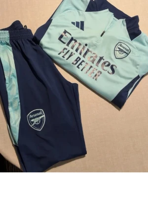 Arsenal tracksuit från adidas - Snyggt Arsenal tröja från Adidas i ljusblått och marinblått. Setet består av långärmad tröja , båda med Arsenal-logga och Adidas-märke. Materialet är funktionspolyester, perfekt för träning. Emirates Fly Better-tryck på tröjan.