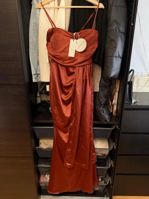 Rostbrun långklänning från Boohoo - Snygg rostbrun långklänning från Boohoo i glansigt satinliknande material. Klänningen har smala axelband, draperad front och en dekorativ ringdetalj vid bysten. Perfekt för dig som vill sticka ut med en elegant look.