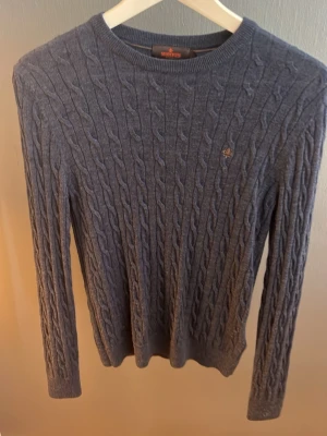 Blå kabelstickad tröja Morris - Kabelstickad blå tröja från Morris med rund halsringning och diskret broderad logga på bröstet. Långärmad modell i mjukt material, perfekt för lager-på-lager. Klassisk och stilren design som funkar till mycket.