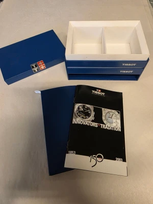 Tissot original klockask och manual och certifikat  - Säljer en blå original klockask från Tissot med vit insida och tillhörande manual. Asken har två fack och Tissot-logga på locket. Perfekt för dig som vill ha en snygg och praktisk förvaring till din Tissot-klocka.