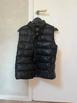 Svart dunväst f - Snygg svart dunväst med hög krage och quiltad design. Västen har dragkedja framtill och är tillverkad i glansigt material som ger en modern look. Perfekt att slänga över en hoodie eller tröja för extra värme.