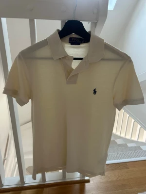 Beige pikétröja från Ralph Lauren - Klassisk vit pikétröja från Polo Ralph Lauren med korta ärmar och blå broderad logga på bröstet. Tröjan har krage, knappar framtill. Perfekt till en clean och stilren look. Säljer den eftersom att den tyvärr inte passar mig längre. Använd mindre än 7 gånger eftersom att jag köpte den förra sommaren vid slutet sen dess har jag växt och den passar inte längre. Tröjan är som helt ny och har inga skador och så vidare. I affär ligger de runt 1000 kr säljer den för 500 kan gå ner vid snabb affär. St S