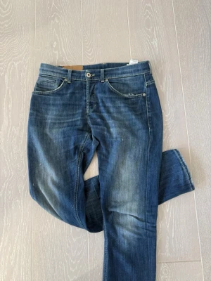 Blå raka jeans med slitningar dondups  - Säljer ett par klassiska blå jeans med raka ben och snygga slitningar framtill. Jeansen har fem fickor, orangea sömmar och en liten logga bak i midjan. Perfekta för en avslappnad och trendig look.