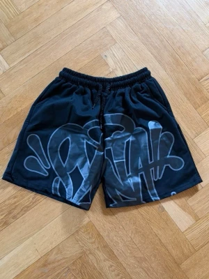 Svarta shorts med grafiskt tryck - Coola svarta shorts med elastisk midja och stort grafiskt tryck i grått och vitt framtill. Perfekta för en avslappnad streetwear-look. Sköna och lätta att bära, passar till många olika outfits.