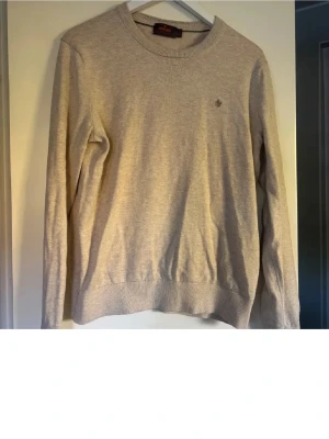 Beige stickad tröja från Gant - Klassisk beige stickad tröja från Gant med rund halsringning och diskret logga broderad på bröstet. Tröjan har långa ärmar och ribbade muddar vid ärmslut och nederkant. Perfekt för lager-på-lager och enkel att matcha.