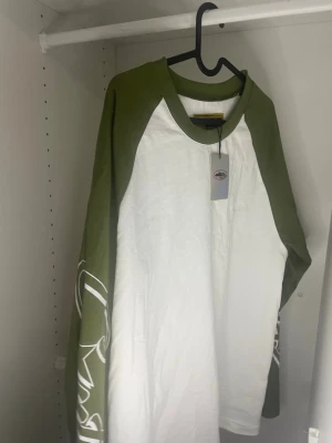 Cortiez långärmad t shirt - Säljer en långärmad t-shirt från Cortiez med vita ärmar och kropp samt gröna kontrastärmar och krage. Tryck i vitt på ena ärmen ger en cool detalj. Perfekt för dig som gillar streetwear och avslappnad stil.
