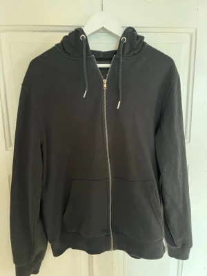 Svart zip-hoodie från 157 - Svart hoodie med dragkedja från 157. Klassisk modell med huva och dragsnören, samt två stora fickor framtill. Tillverkad i mjukt material som är skönt att ha på sig. Perfekt för en avslappnad stil.