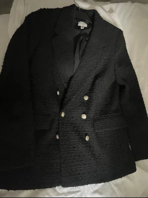 Svart kavaj med guldknappar - Stilren svart kavaj med dubbelknäppning och sex dekorativa guldknappar framtill. Kavajen har en strukturerad yta och två fickor. Perfekt för en trendig och klassisk look.