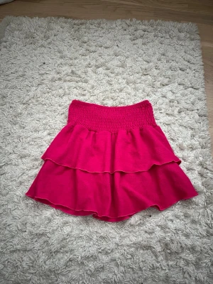 Rosa volangkjol med smock - Superfin rosa kjol med bred smock i midjan och dubbla volanger. Kjolen har en kort och lekfull modell som passar perfekt till varma dagar. Materialet känns mjukt och följsamt.