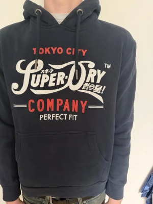 Mörkblå Superdry hoodie med tryck - Snygg mörkblå hoodie från Superdry med stort vitt och rött tryck på bröstet. Klassisk känguruficka framtill, dragsko i huvan och mjukt material. Perfekt för en avslappnad streetstyle-look.