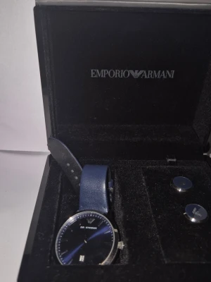 Emporio Armani blå klocka - Stilren klocka från Emporio Armani med blått läderarmband och blå urtavla. Klockan har silverfärgad boett och detaljer samt datumvisning. Perfekt accessoar för dig som gillar klassisk och modern design.