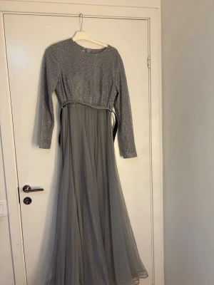 Lång grå klänning med glitterdetaljer - Elegant långklänning i grått med glittrig överdel och långärmade ärmar. Kjolen är i ett tunt, svepande material och midjan markeras med ett smalt bälte. Perfekt för speciella tillfällen när du vill sticka ut.