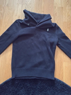 Svart stickad tröja Ralph Lauren Sport - Svart stickad tröja från Ralph Lauren Sport med stor krage och knappdetalj. Tröjan har lång ärm, ribbstickade muddar och den klassiska vita logotypen broderad på bröstet. Tillverkad i 100% bomull för en mjuk och bekväm känsla. Öppen för pris förslag