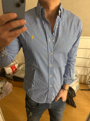Blårandig skjorta från Ralph Lauren - Säljer en klassisk blårandig skjorta från Ralph Lauren med vit knappstängning och gul broderad logga på bröstet. Skjortan har lång ärm, button-down krage och är gjord i mjuk bomull. Perfekt för en clean och stilren look.