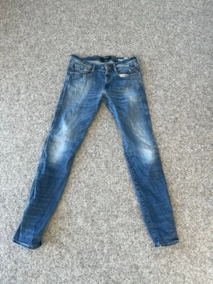 Blå slim fit jeans med slitningar - Säljer ett par blå slim fit jeans med snygga slitningar och tvättade detaljer. Klassisk femficksmodell med dragkedja och knapp i midjan. Jeansen har en smal passform och är tillverkade i denim, perfekt för en avslappnad och trendig look. Köptes för 1599kr men säljs för 159kr på grund av slitningar 