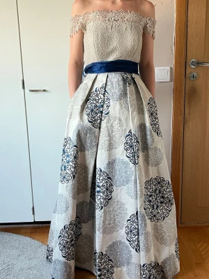 Offshoulder långklänning med spets - Elegant offshoulder långklänning med spetsliv och vid kjol i vitt och blått mönster. Midjan markeras av ett brett, mörkblått band. Kjolen har stora dekorativa mönster i blått och grått och klänningen har fickor. Perfekt för bal eller festliga tillfällen. Klänningen är insydd o h uppsydd till en 32 men går att sprätta upp till en 34