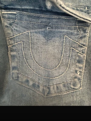 Ljusblå bootcut jeans från True Religion - Säljer ett par ljusblå jeans från True Religion med ikonisk vit kontrastsöm och broderad bakficka. Jeansen har bootcut-modell och klassisk femficksdesign. Perfekta för dig som gillar en avslappnad men trendig look. De är bra skick men slitningar längst ner på benen och en knapp har även försvunnit på en bak ficka.🩷säljer inte för under 350