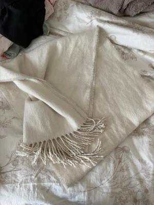 Beige halsduk med fransar - Mjuk och stilren beige halsduk med dekorativa fransar i ändarna. Halsduken har en rektangulär form och är tillverkad i ett skönt material som känns lyxigt mot huden. Perfekt för att addera en enkel men snygg detalj till din outfit.