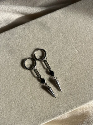 Silvriga örhängen med svarta detaljer - Säljer ett par coola silvriga örhängen med ring och hängande stav samt svart detalj och spetsig kon längst ner. Örhängena har en edgy och modern design som sticker ut. Perfekta för dig som gillar statement-smycken.