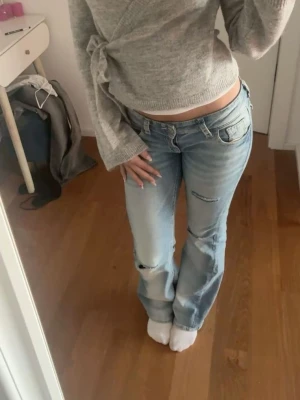 Ljusblå bootcut jeans med slitningar - Säljer ett par ljusblå bootcut jeans med slitningar på knäna och lätt tvättad look. Jeansen har klassisk femficksdesign och låg midja. Perfekta för en avslappnad och trendig stil.
