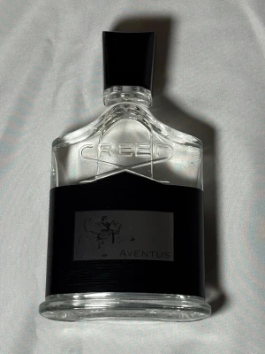 Creed Aventus Eau de Parfum 100ml - Creed Aventus är en exklusiv parfym i en elegant, transparent glasflaska med svart etikett och svart plastkork. Flaskan har en stilren design med märkets logotyp ingraverad och motiv av en ryttare på etiketten. Volymen är 100 ml.