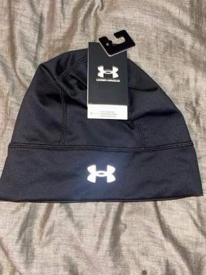Svart mössa från Under Armour - Snygg svart mössa från Under Armour med reflekterande logga framtill. Tillverkad i ett stretchigt funktionsmaterial som passar perfekt för träning eller kalla dagar. Enkel design med synliga sömmar och bekväm passform.