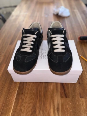 Svarta sneakers från Maison Margiela - Maison Margiela sneakers i svart läder och mocka med vita snören och beige gummisula. Klassisk low-top modell med stilren design och diskreta detaljer. Perfekt för dig som vill ha en clean och tidlös look. Skick 7/10. Pris ej hugget i sten.