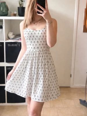 Vit blommig kortklänning - Söt vit kortklänning med små svarta rosor, smal passform upptill och utställd kjol. Klänningen är ärmlös med smala axelband och har en feminin, somrig look. Perfekt för varma dagar och enkel att styla med smycken eller en jeansjacka.