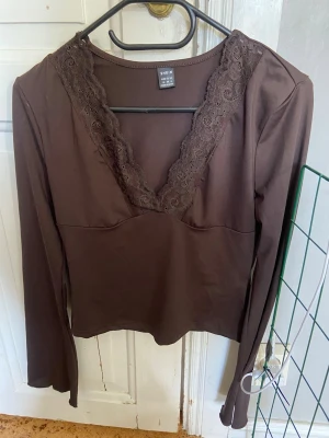 Brun långärmad topp med spets SHEIN - Mörkbrun långärmad topp från SHEIN med djup v-ringning dekorerad med spetsdetaljer. Tillverkad i mjukt material som sitter bekvämt och har en feminin touch med spetsen runt halsen. Perfekt att styla med jeans eller kjol. Oanvänd